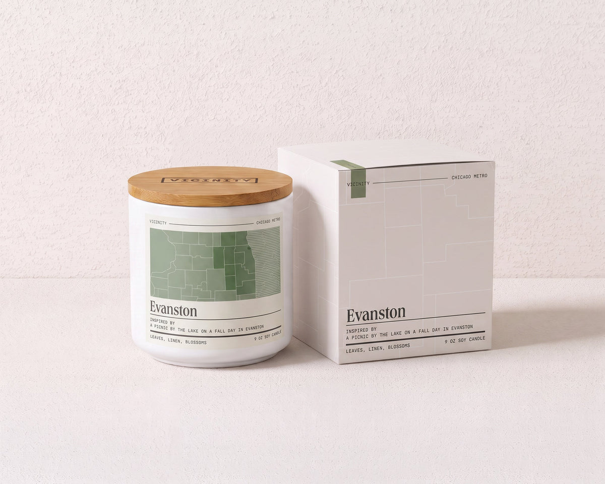 Evanston 9 oz soy candle and its gift box, both printed with a Chicago neighborhood map, on a cream plaster surface